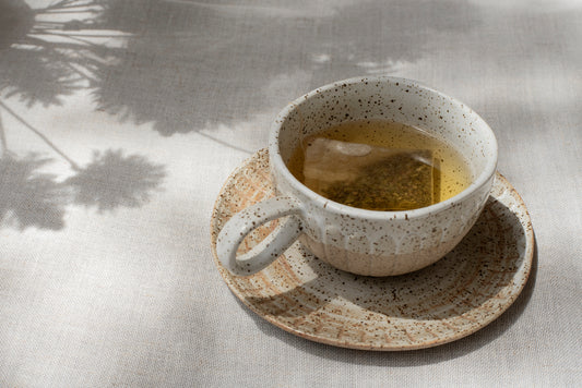 Angled overhead showing pottery-style cup and saucer in shades of brown and beige with tea and teabag inside sitting on a grey-beige linen cloth with shadows of flowers cast nearby