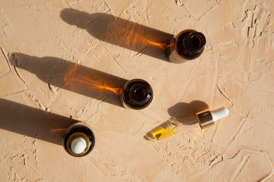 Three amber tincture bottles shown from overhead on terra-cotta surface with one dropper out and laying on it with sunlight creating slanted shadows from the bottles